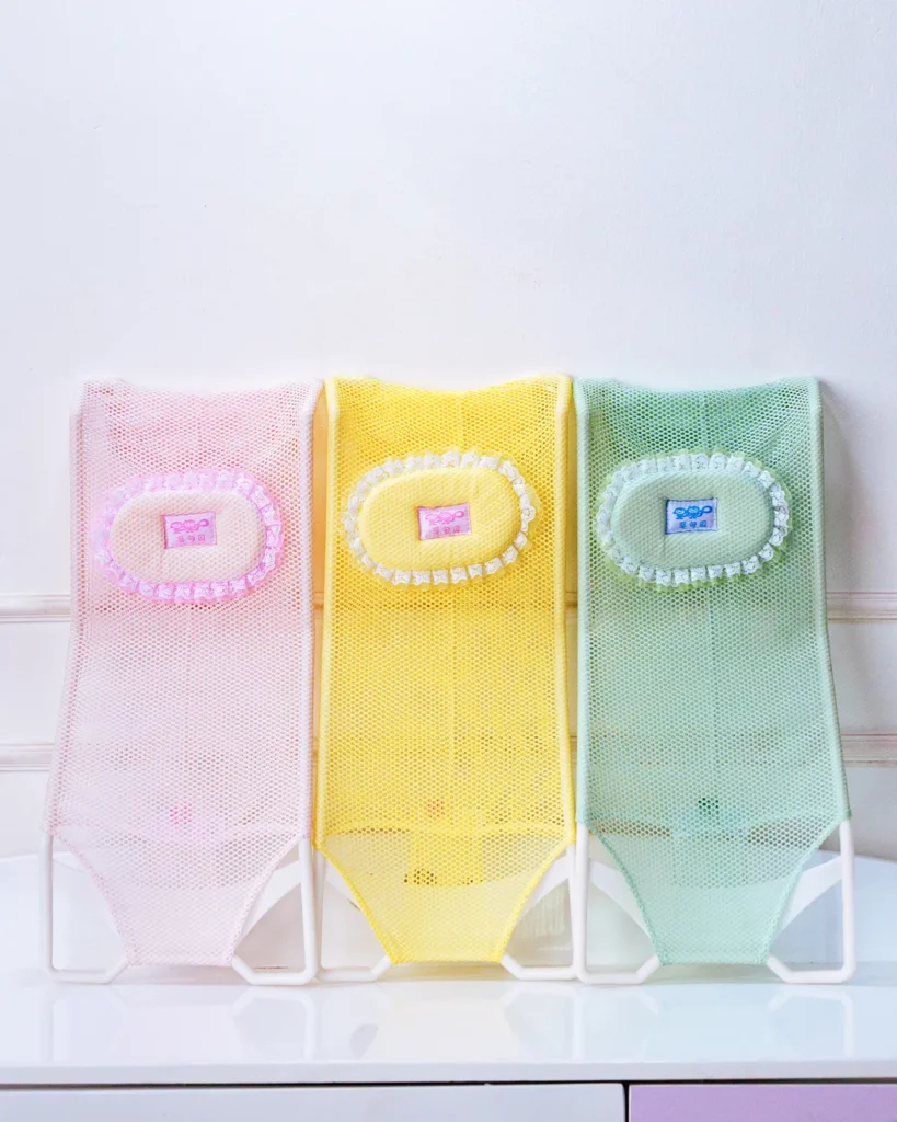 Group shot of three colorful baby bath support net slings (pink, yellow, green) side-by-side.