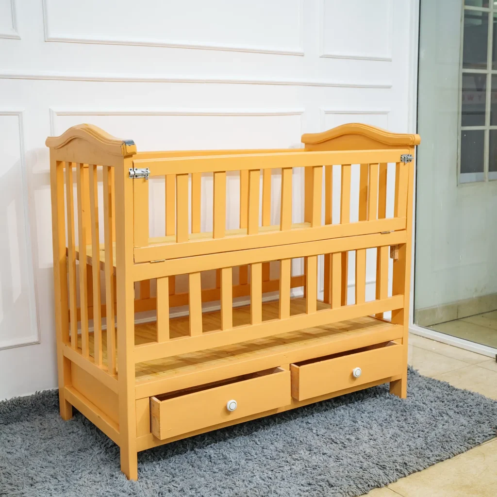 Angled view of a natural wood finish baby cot bed with two storage drawers (white knobs) and visible hinges suggesting an adjustable side rail, placed on a gray shag carpet near a white wall and doorway.