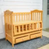 Wooden Baby Cot Bed 5 A Natural Wood Finish Baby Cot With Two Drawers, Viewed From An Angle On A Gray Carpet Near A White Wall.