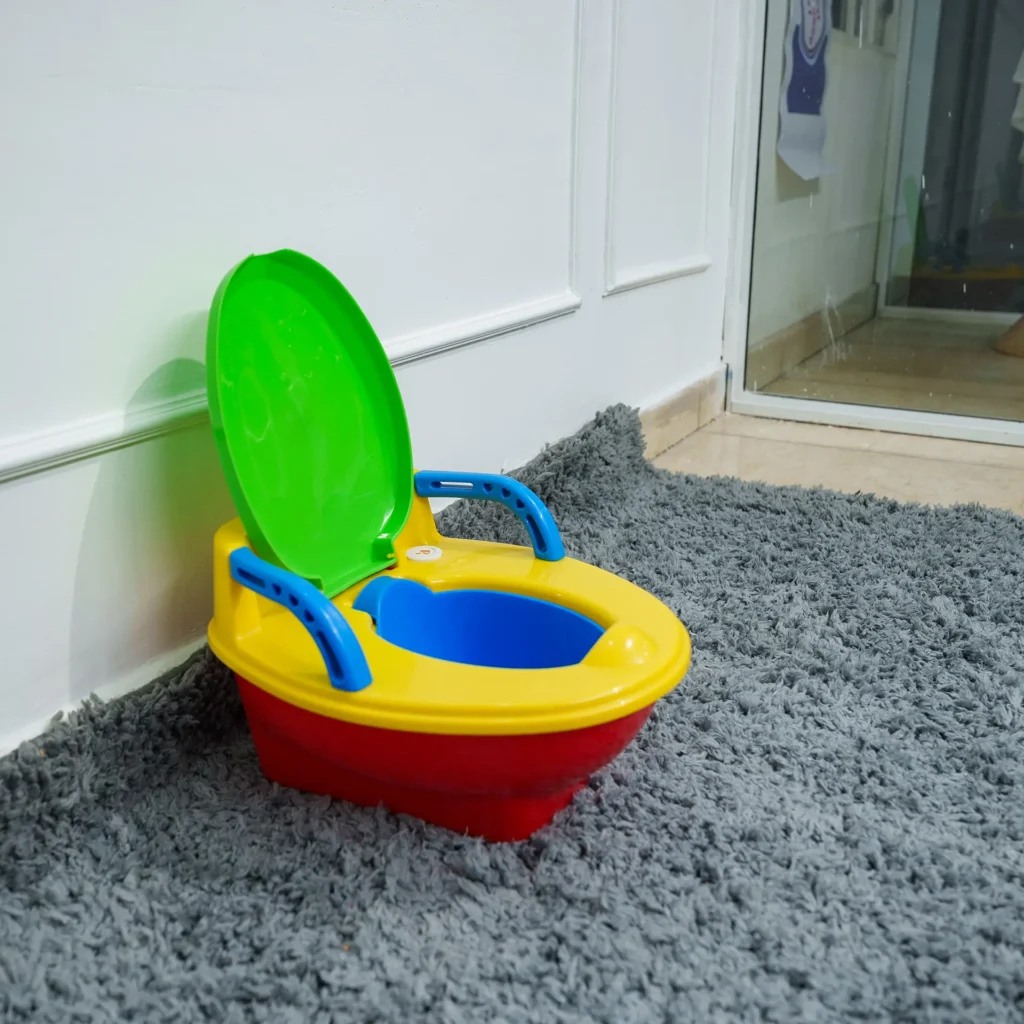 Angled view of the colorful child's potty chair (red base, yellow seat, blue handles, open green lid) sitting on a gray shag carpet near a white wall and a glimpse of a doorway.