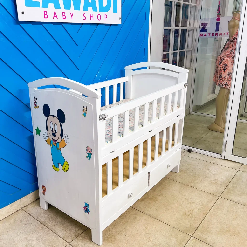 Front view of a white wooden baby cot with mattress and storage drawers at Zawadi Baby Shop
