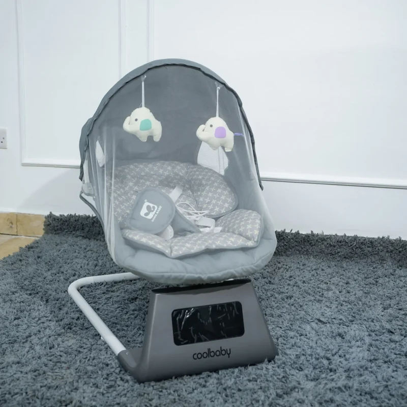 Front view of the grey CoolBaby electric baby swing, highlighting the padded seat with pattern, canopy with attached soft toys, protective mosquito net, and control base, set on a grey rug.