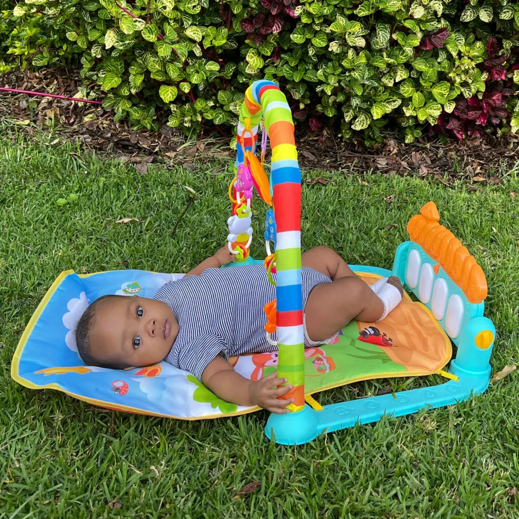 A colorful Piano Play Mat for Toddlers featuring a baby lying on a cushioned mat with hanging toys and a touch-sensitive piano, set outdoors on green grass.