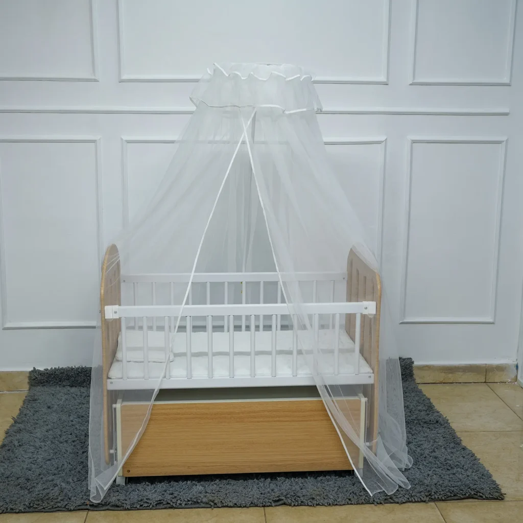 Front view of a wooden baby crib featuring white railings, a canopy mosquito net fully draped, and an under-crib storage drawer, placed on a grey rug.