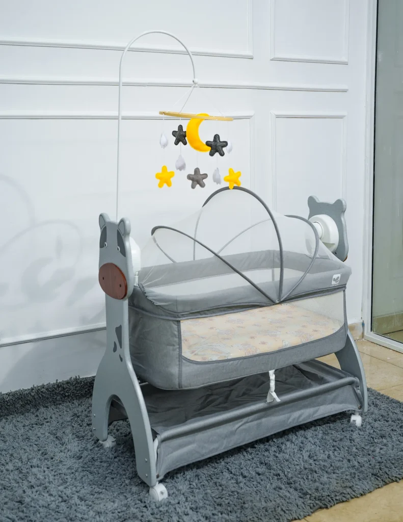 Angled side view of a grey electronic newborn bassinet featuring an animal-themed frame, mesh sides, mosquito net canopy, overhead mobile with stars and moon, and lower storage shelf, placed on a grey rug.