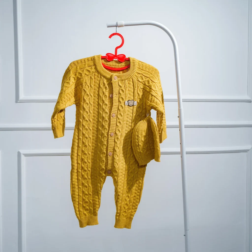 Mustard yellow cable knit baby jumpsuit with button front and matching hat, hanging on a white stand against a white paneled wall