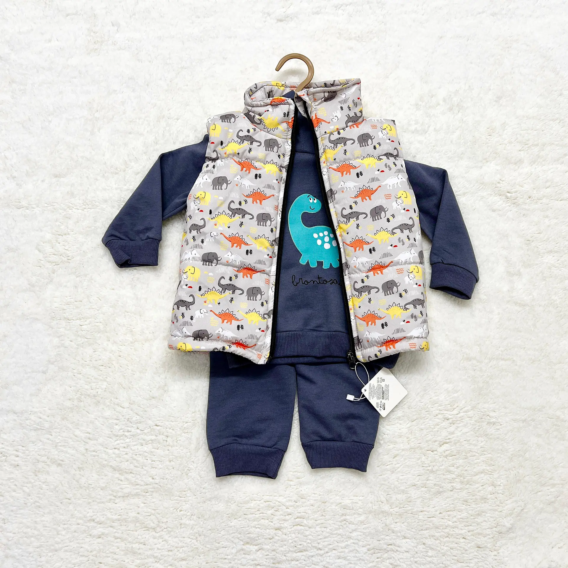 3 Pcs Baby Outfits 4 A 3-Piece Baby Outfit Set Displayed On A Cream Background, Including A Dark Blue Long-Sleeve Sweatshirt With A Teal Brontosaurus Graphic, Matching Pants, And A Grey Sleeveless Puffer Vest With Multicolor Dinosaur Prints.