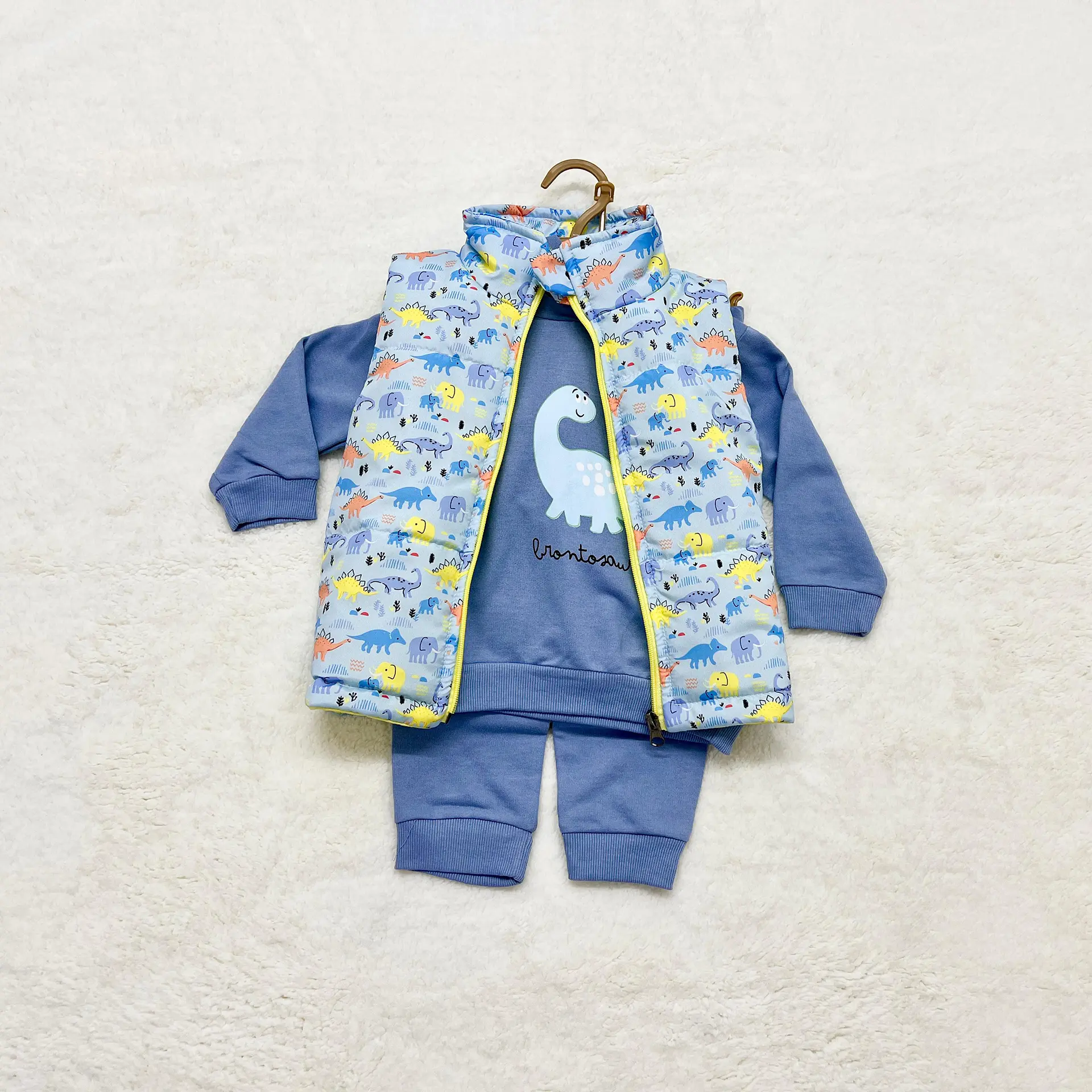3 Pcs Baby Outfits 3 A 3-Piece Baby Outfit Set Laid Flat On A Soft Cream Surface, Featuring A Light Blue Long-Sleeve Sweatshirt With A Cartoon Brontosaurus, Matching Pants, And A Sleeveless Puffer Vest With Colorful Dinosaur Prints.