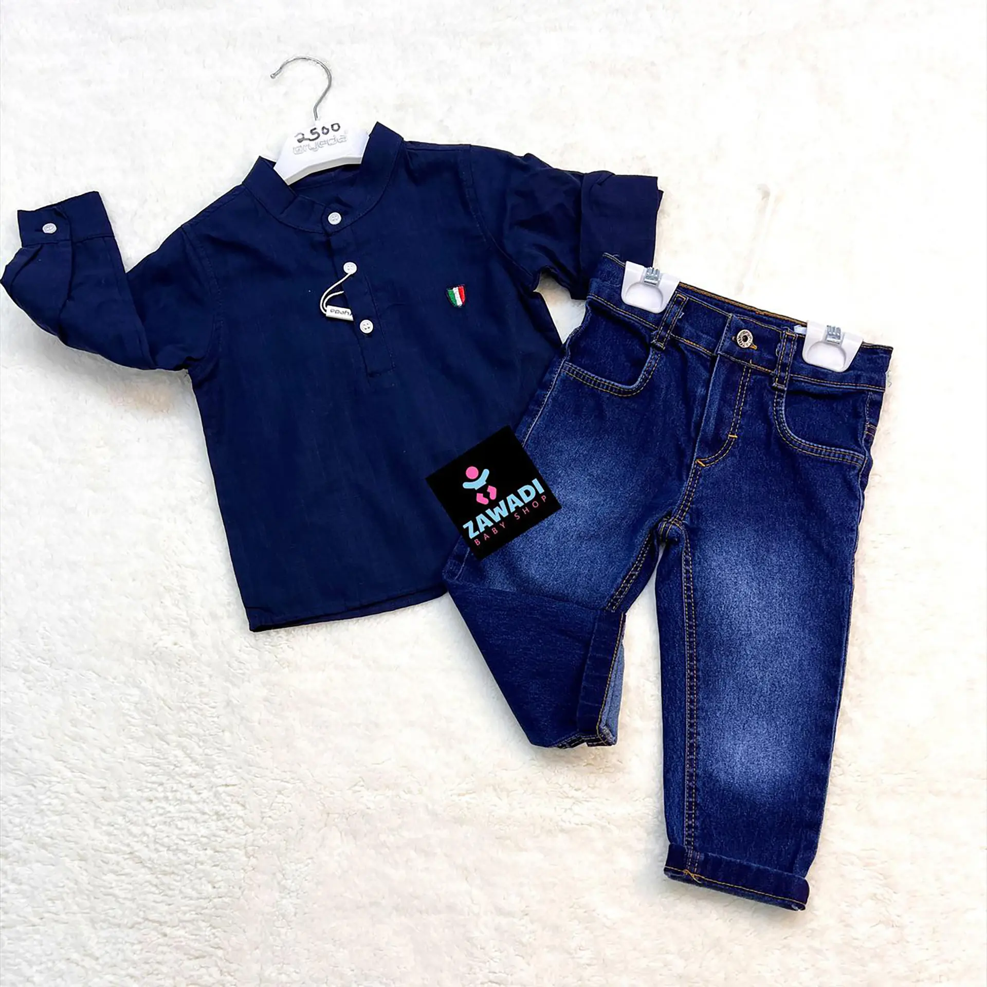 Boys Jeans Set 2 Flat Lay Of A Boys' Outfit Featuring A Long-Sleeved Dark Blue Shirt With A Small Embroidered Patch And A Pair Of Classic Blue Jeans, Displayed On A White Textured Background. The Set Is Labeled With A Zawadi Baby Shop Tag.