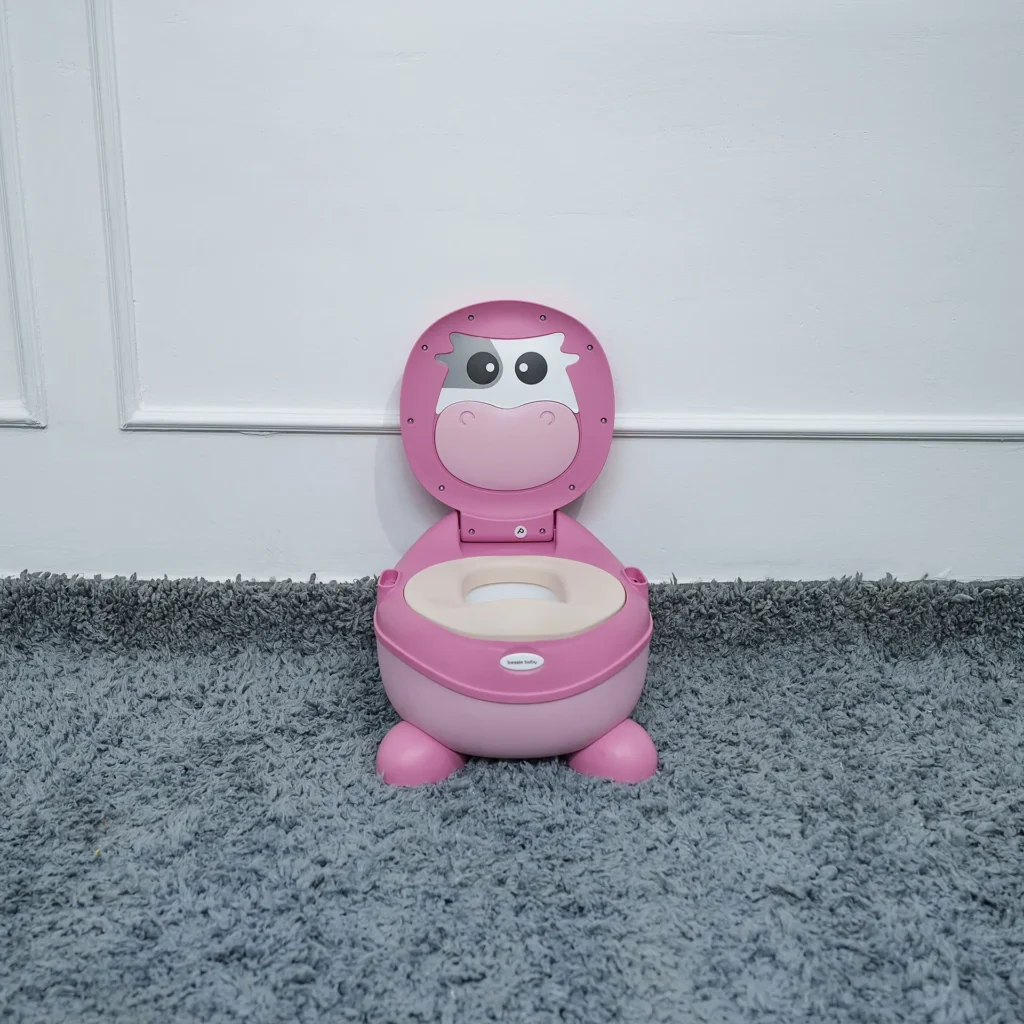 Pink and white toddler potty with a cartoon cow face on the lid, placed on a gray carpet in front of a white wall.