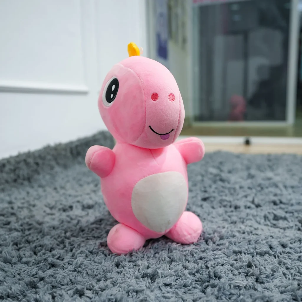 Pink plush soft toy with a white belly and friendly face, displayed on a grey carpet in a nursery setting – ideal for babies and toddlers.