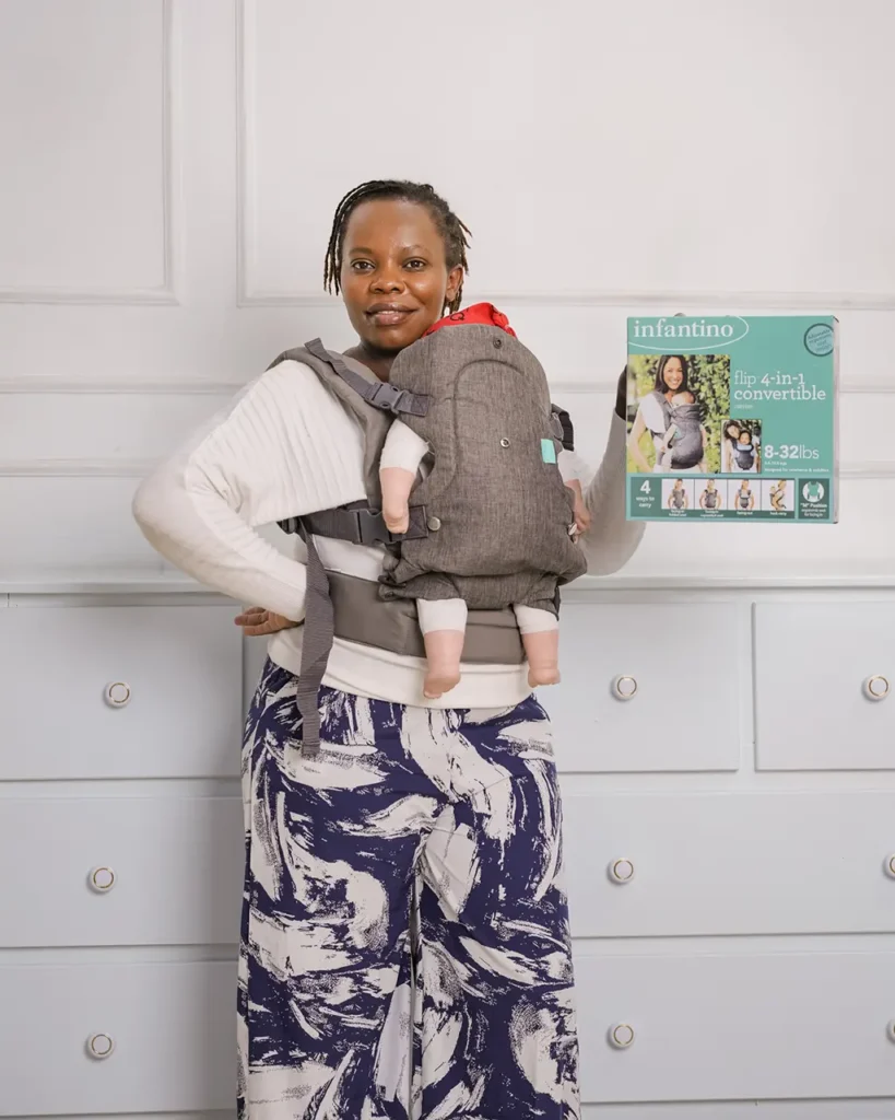 Kui Thairu wearing the Infantino 4-in-1 baby carrier facing in, holding its box, at Zawadi Baby Shop Nairobi.
