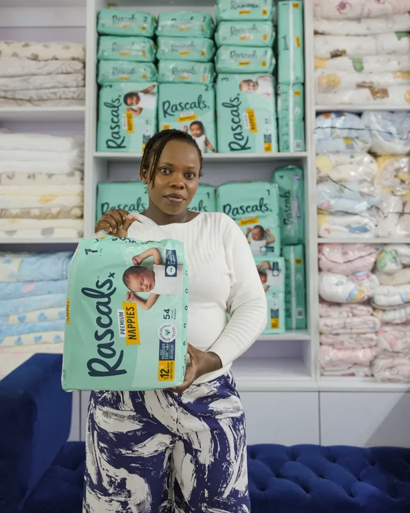 Kui Thairu holding a Size 1 pack of Rascals diapers beside shelves of Rascals stock at Zawadi Baby Shop Nairobi.